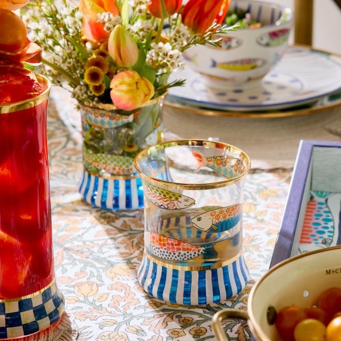 The Kitchen Table Go Fish highball glass with hand-painted fish, navy stripes, and gold lustre rim on a floral table
