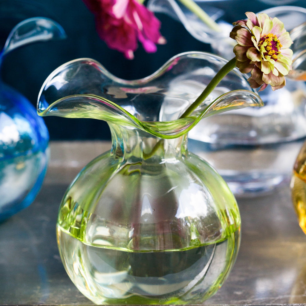The Kitchen Table Hibiscus glass fluted bud vase in clear light green with a delicate ruffled rim holding a flower.