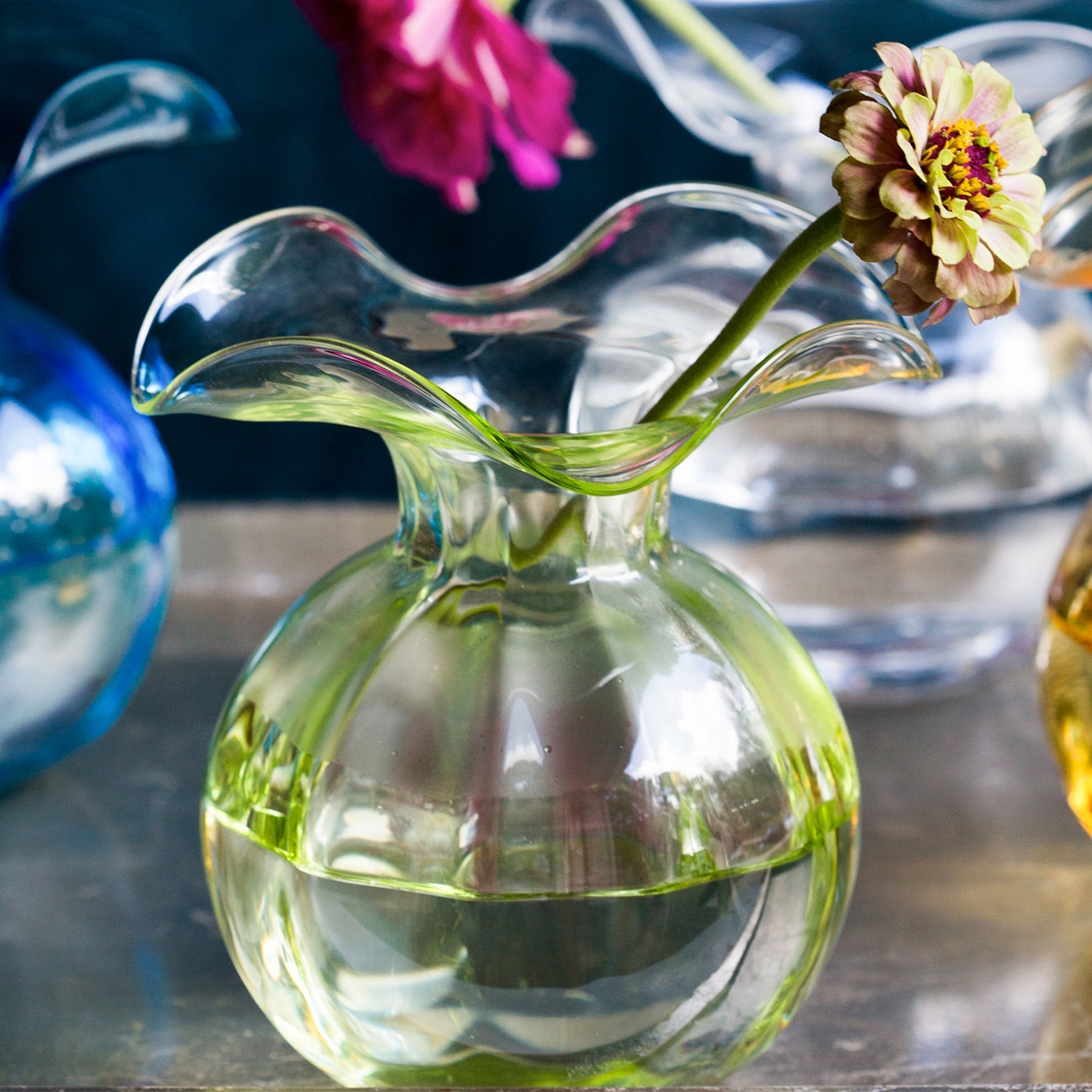 The Kitchen Table Hibiscus glass fluted bud vase in clear light green with a delicate ruffled rim holding a flower.