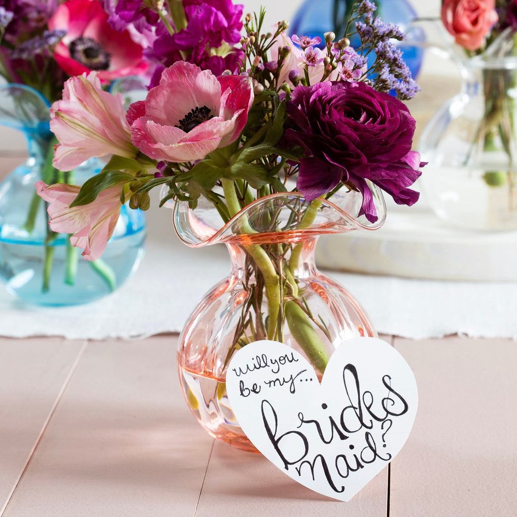 The Kitchen Table Hibiscus glass fluted bud vase in pink holding a floral arrangement with a bridesmaid heart tag.