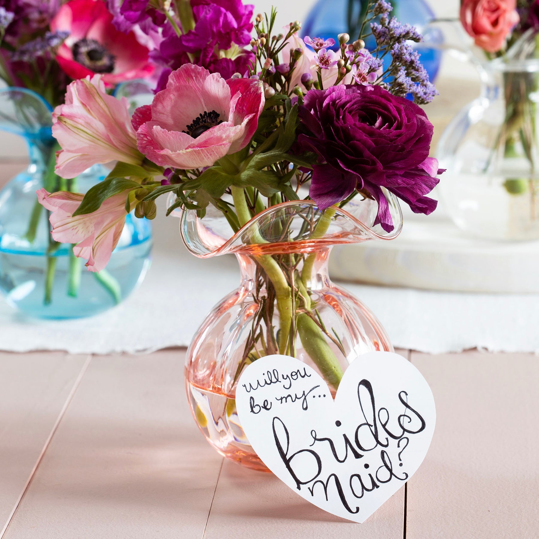 The Kitchen Table Hibiscus glass fluted bud vase in pink holding a floral arrangement with a bridesmaid heart tag.