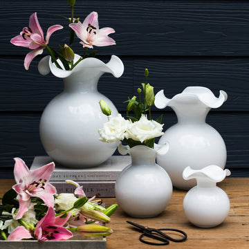 The Kitchen Table Hibiscus glass fluted vases in white with ruffled rims, displayed with fresh flowers on a wooden table.