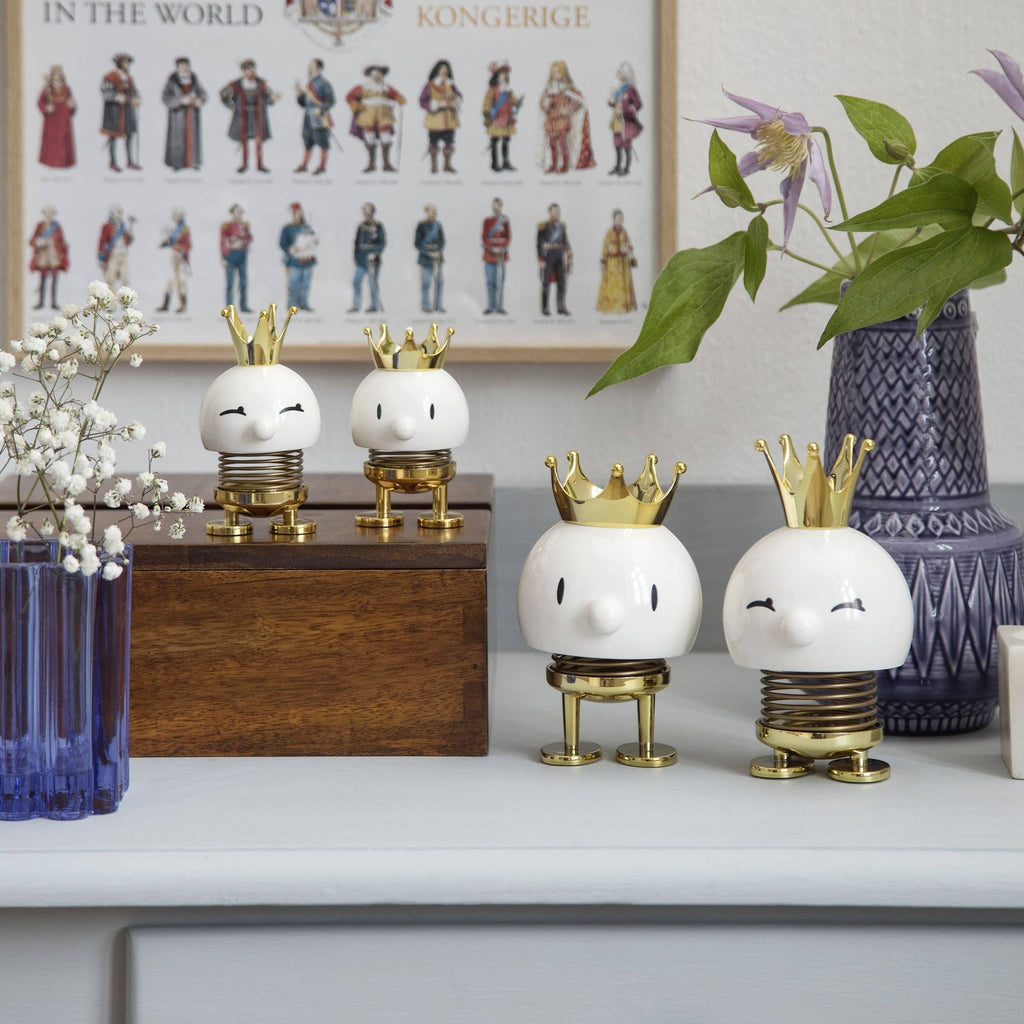 The Kitchen Table Hoptimist Queen figurines with white bodies, gold crowns, and spring legs on a shelf.