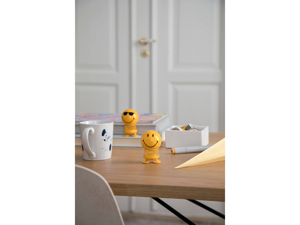 The Kitchen Table Hoptimist Smiley S yellow spring figurine with a beaming smile on a wooden desk