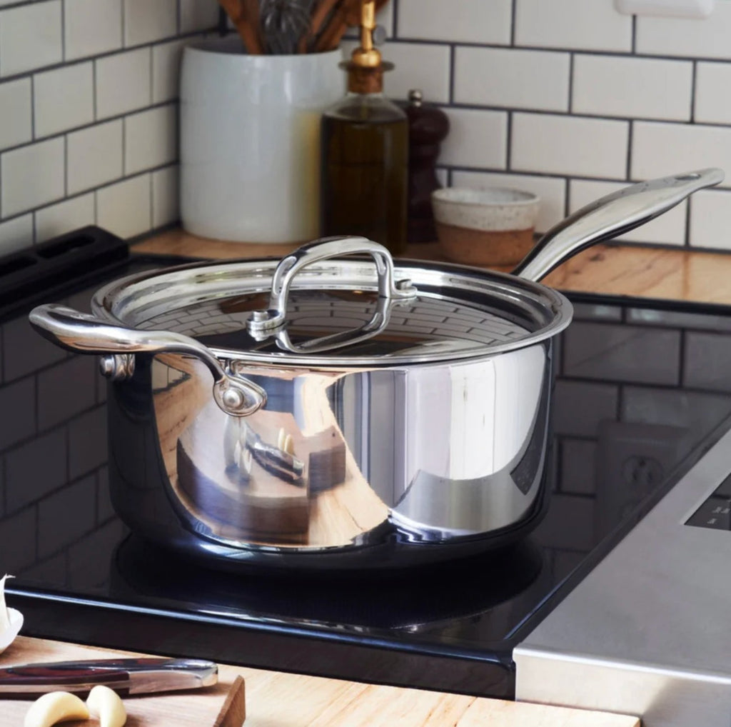 The Kitchen Table 4 quart stainless steel saucepan with lid on a black stovetop in a modern kitchen.