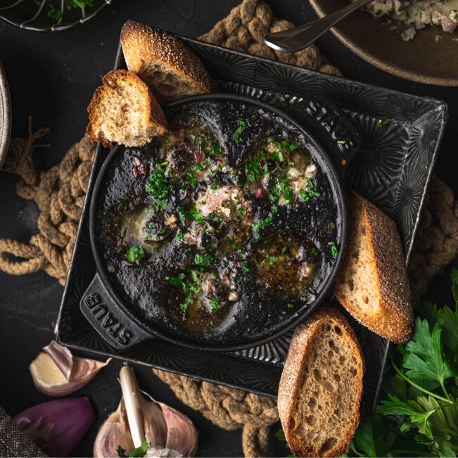 The Kitchen Table Staub cast iron escargot dish filled with snails in herb butter, served with crusty bread.