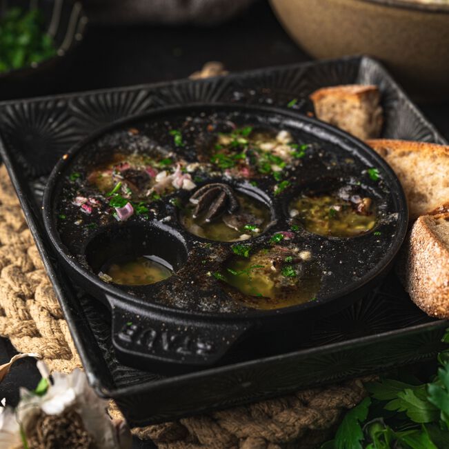 The Kitchen Table Staub cast iron escargot dish with six holes, filled with prepared snails in herb butter.